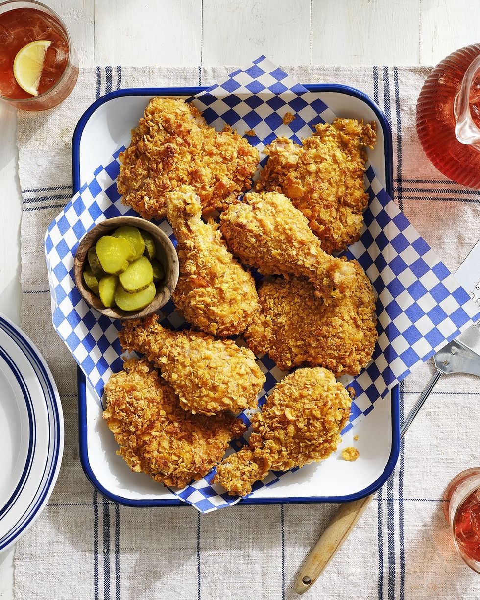 cornflake crusted baked chicken on a white tray lined with blue and white checkered parchment paper and a small bowl of sliced pickles